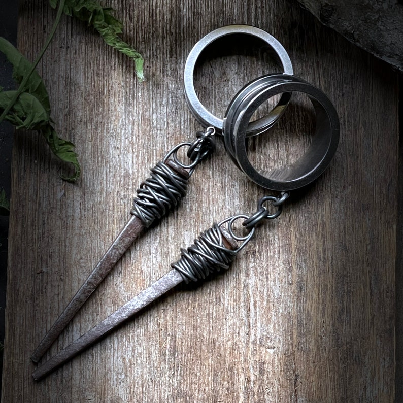 May include: A silver key chain with two metal rings and two wire-wrapped nails. The nails are attached to the rings with a silver chain.