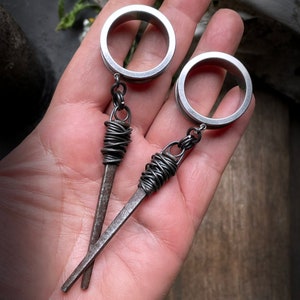 May include: Two silver metal hoop earrings with a dangling wire wrapped with brown thread and a pointed tip. The earrings are hanging from a hand.