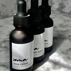 Puede incluir: Tres botellas de vidrio con goteros negros y etiquetas blancas. Las etiquetas tienen texto negro que dice "soma ROSE SERUM ULTRA HYDRATING FACIAL OIL", "DER SERUM ALING FACIAL OIL", y "RUM TENING FACIAL OIL".