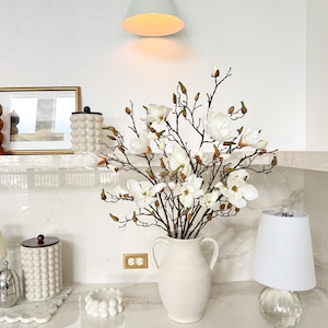 May include: A white ceramic vase with two handles holds a bouquet of white magnolia flowers and buds. A white lamp with a clear base and a white lampshade sits to the right. A white wall-mounted light fixture is above the vase.