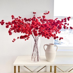May include: A clear glass vase holds bright red floral branches. A textured beige ceramic pitcher with two handles sits on a white table with gold legs. The scene is set against a white wall and a window, creating a simple, elegant display.