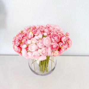 May include: A bouquet of pink and white ranunculus flowers in a clear glass vase with a gold metal frame.