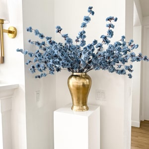 May include: A metallic gold vase with a smooth, rounded body and a flared base, filled with numerous branching stems of small, light blue artificial flowers that extend outwards in a wide, airy arrangement against a plain white wall.
