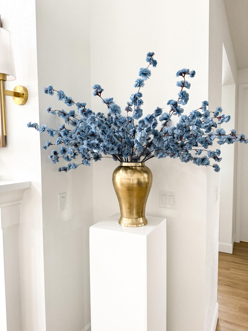 May include: A metallic gold vase with a smooth, rounded body and a flared base, filled with numerous branching stems of small, light blue artificial flowers that extend outwards in a wide, airy arrangement against a plain white wall.