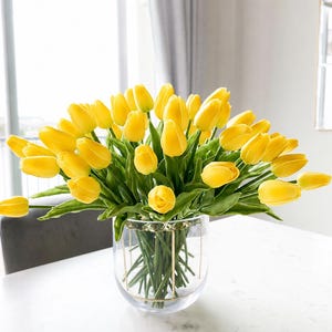 Puede incluir: Un gran ramo de tulipanes de color amarillo vibrante con tallos y hojas verdes dispuestos en un jarr&oacute;n de cristal transparente. El jarr&oacute;n presenta una fina estructura de alambre met&aacute;lico dorado en el exterior que sostiene los tallos.