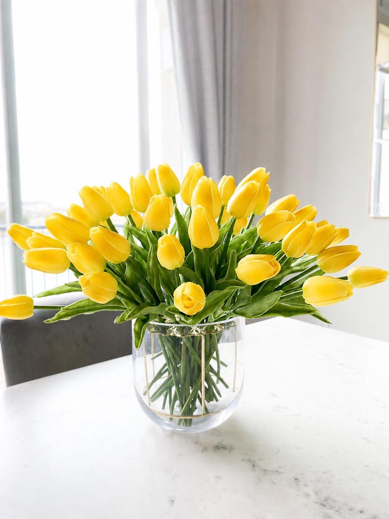 Puede incluir: Un gran ramo de tulipanes de color amarillo vibrante con tallos y hojas verdes dispuestos en un jarr&oacute;n de cristal transparente. El jarr&oacute;n presenta una fina estructura de alambre met&aacute;lico dorado en el exterior que sostiene los tallos.