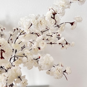 May include: A cluster of artificial cherry blossom branches featuring numerous small, layered white flowers attached to thin, dark brown stems. The delicate petals are arranged in dense groupings along the branching structure, creating a full appearance.