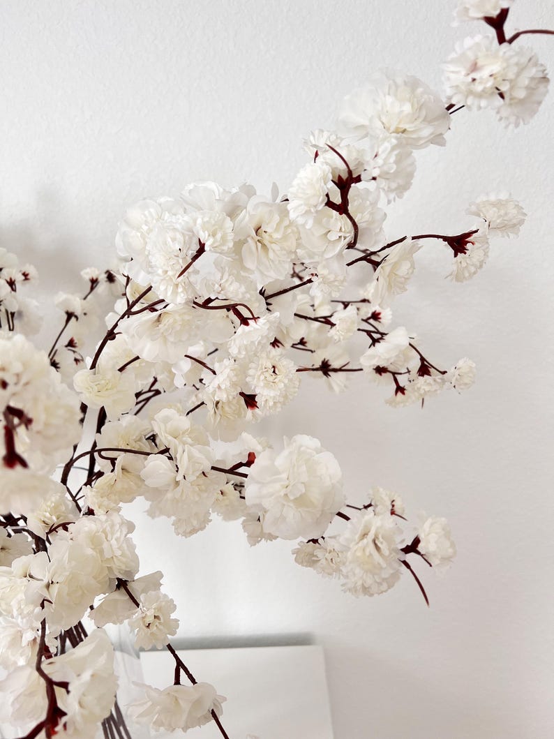 May include: A cluster of artificial cherry blossom branches featuring numerous small, layered white flowers attached to thin, dark brown stems. The delicate petals are arranged in dense groupings along the branching structure, creating a full appearance.