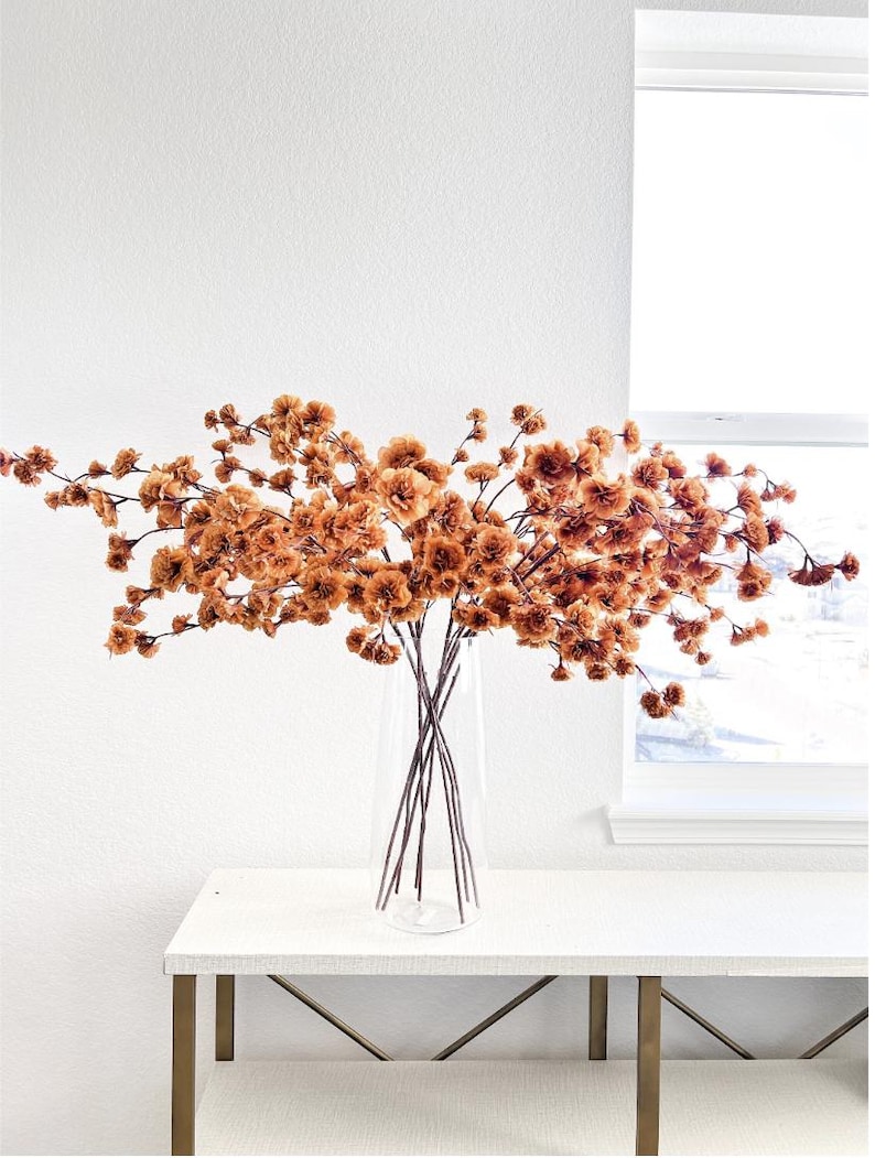 May include: A clear glass vase containing multiple stems of dried, rust-coloured flowers. The branches spread horizontally, featuring numerous small, ruffled blossoms in shades of burnt orange and brown, creating a wide, airy arrangement.