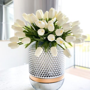 Puede incluir: Un gran ramo de tulipanes blancos con tallos y hojas verdes dispuesto en un jarr&oacute;n cil&iacute;ndrico de cristal. El jarr&oacute;n presenta una secci&oacute;n superior texturizada con patr&oacute;n de diamantes, una banda lisa de color cobre y una base plateada reflectante.