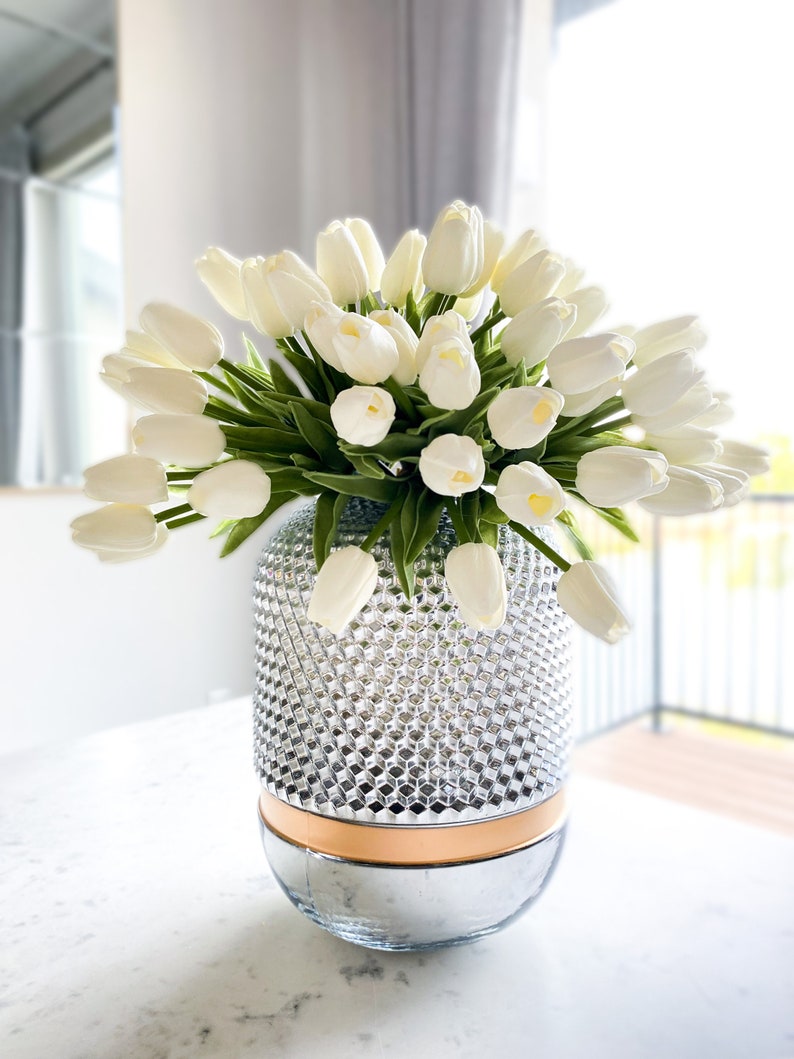 Puede incluir: Un gran ramo de tulipanes blancos con tallos y hojas verdes dispuesto en un jarr&oacute;n cil&iacute;ndrico de cristal. El jarr&oacute;n presenta una secci&oacute;n superior texturizada con patr&oacute;n de diamantes, una banda lisa de color cobre y una base plateada reflectante.