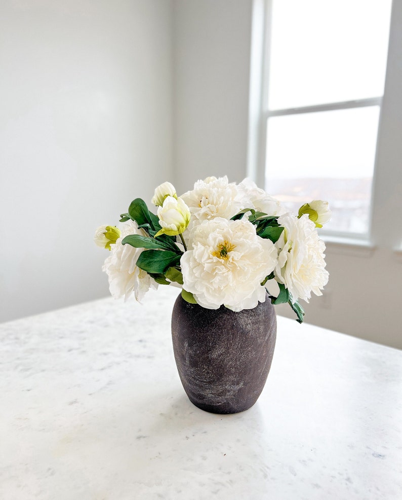 May include: A bouquet of white peonies in a dark gray ceramic vase on a white marble countertop.