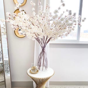 May include: A tall, clear glass vase filled with numerous artificial white cherry blossom branches. The vase sits on a round white marble table with a gold metal base. A small, clear glass decorative orb rests on a gold beaded tray on the table.