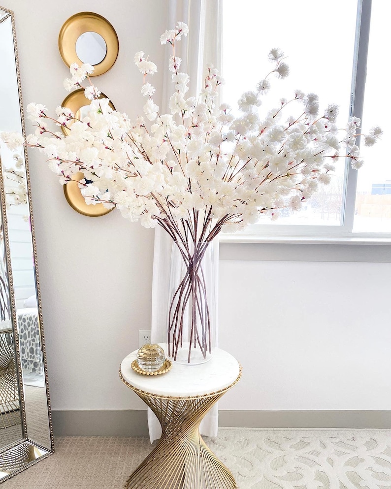 May include: A tall, clear glass vase filled with numerous artificial white cherry blossom branches. The vase sits on a round white marble table with a gold metal base. A small, clear glass decorative orb rests on a gold beaded tray on the table.