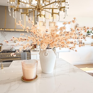 May include: A white ceramic vase with two side handles filled with light peach-coloured faux cherry blossom branches, positioned next to a textured glass candle in a gold-rimmed holder on a white marble surface with grey veining.
