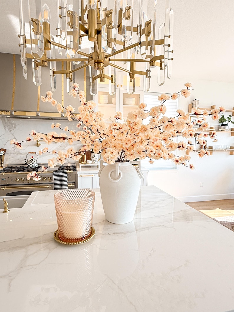 May include: A white ceramic vase with two side handles filled with light peach-coloured faux cherry blossom branches, positioned next to a textured glass candle in a gold-rimmed holder on a white marble surface with grey veining.