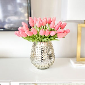 Puede incluir: Un ramo de tulipanes de color rosa suave con tallos y hojas verdes dispuesto en un jarr&oacute;n redondo de vidrio mercurio plateado, con una superficie texturizada y con hoyuelos que refleja la luz en su cuerpo exterior met&aacute;lico.
