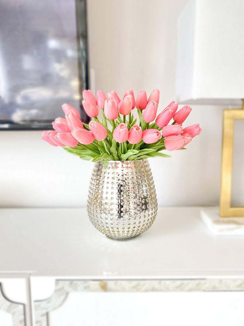 Puede incluir: Un ramo de tulipanes de color rosa suave con tallos y hojas verdes dispuesto en un jarr&oacute;n redondo de vidrio mercurio plateado, con una superficie texturizada y con hoyuelos que refleja la luz en su cuerpo exterior met&aacute;lico.