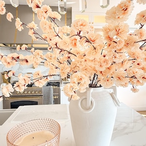 May include: A large white ceramic vase with two ring-shaped handles, filled with numerous branches of pale peach-coloured artificial cherry blossoms. Beside the vase sits a textured, light pink glass candle holder containing a white wax candle.