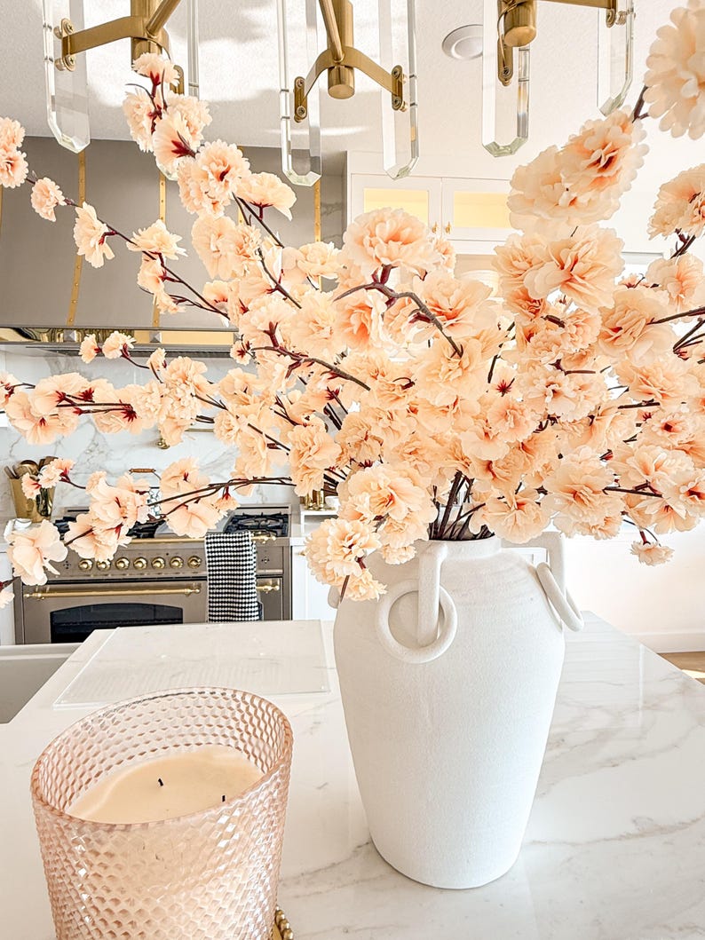 May include: A large white ceramic vase with two ring-shaped handles, filled with numerous branches of pale peach-coloured artificial cherry blossoms. Beside the vase sits a textured, light pink glass candle holder containing a white wax candle.