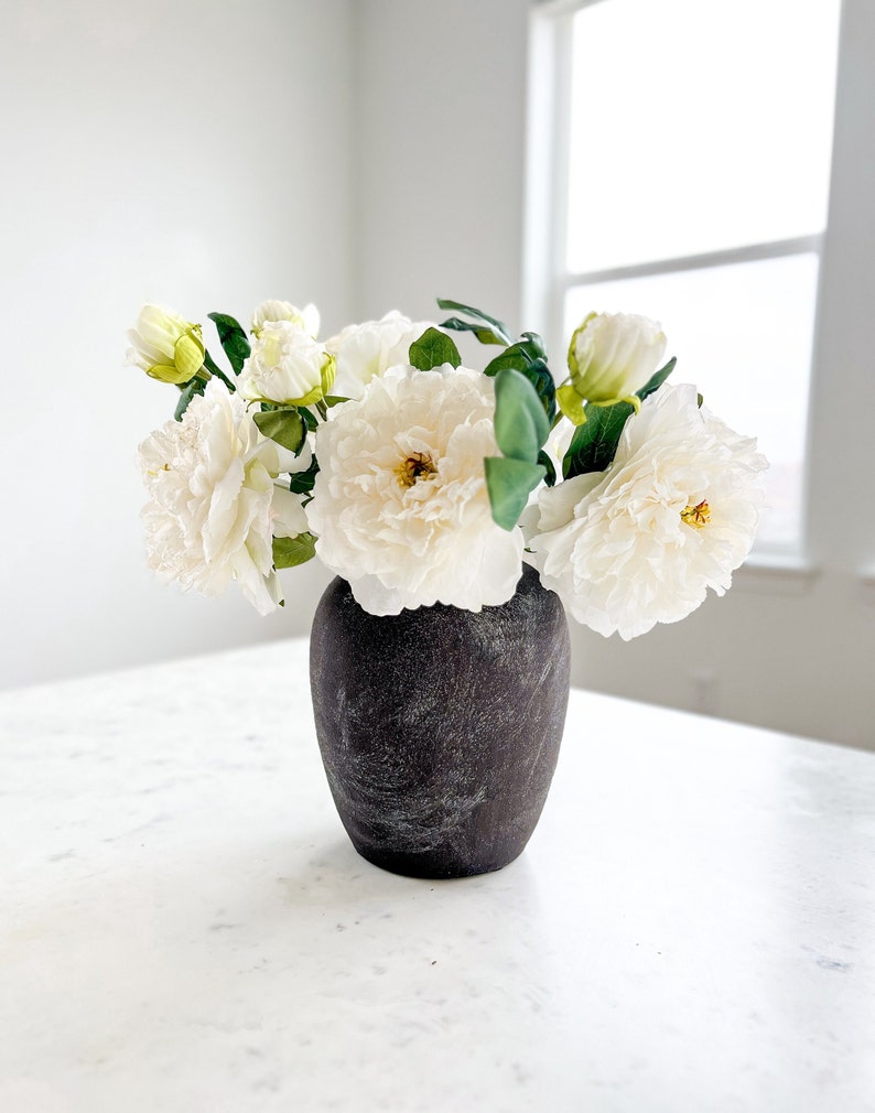 May include: A bouquet of white paper flowers in a dark gray ceramic vase on a white marble surface.