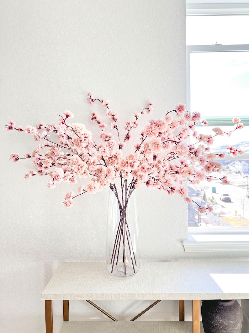 May include: A clear glass vase containing multiple long, slender brown branches adorned with numerous small, pale pink blossoms. The branches spread outward in a wide, fan-like arrangement, creating a dense display of delicate floral clusters.