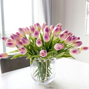 Puede incluir: Un gran ramo de tulipanes artificiales con p&eacute;talos blancos de puntas moradas y tallos verdes, dispuesto en un florero de vidrio transparente con un marco de alambre de metal dorado en el exterior, mostrando m&uacute;ltiples flores en racimo.