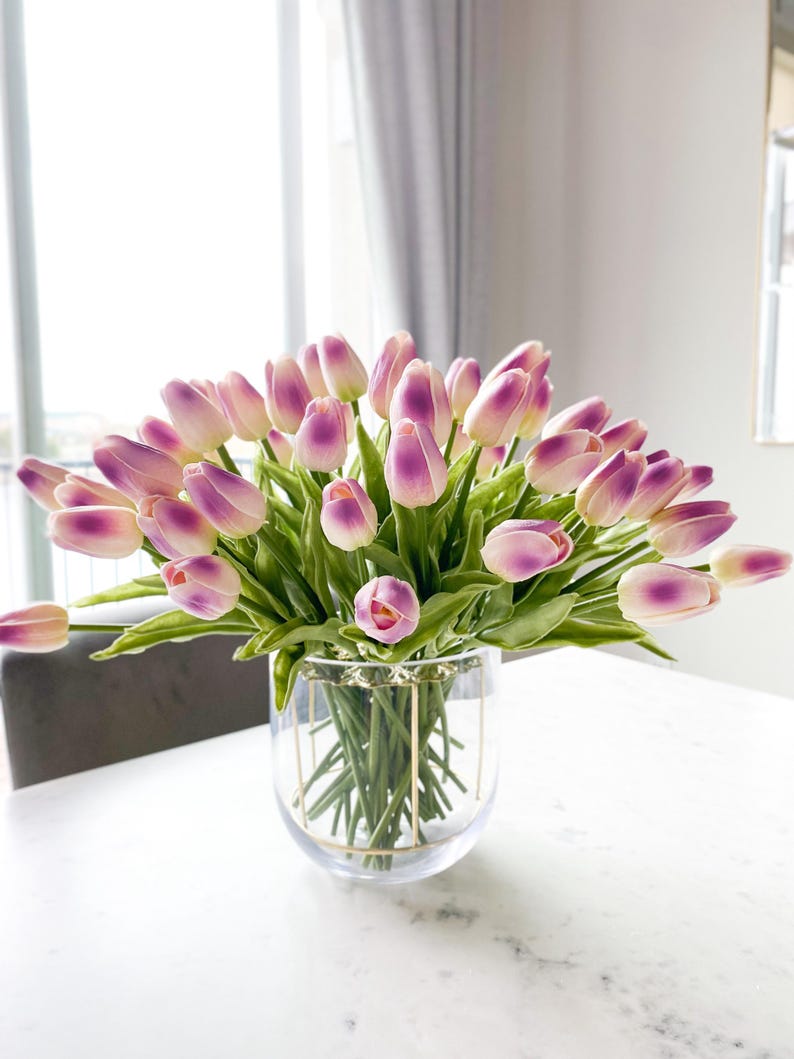 Puede incluir: Un gran ramo de tulipanes artificiales con p&eacute;talos blancos de puntas moradas y tallos verdes, dispuesto en un florero de vidrio transparente con un marco de alambre de metal dorado en el exterior, mostrando m&uacute;ltiples flores en racimo.