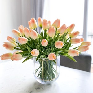 May include: A bouquet of peach-colored artificial tulips arranged in a clear glass vase with a gold metal frame. The vase is sitting on a white table.