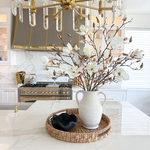 Puede incluir: Una isla de cocina presenta una bandeja tejida con un jarr&oacute;n blanco texturizado lleno de ramas de magnolia blancas. Un cuenco decorativo negro est&aacute; sobre la bandeja. Una l&aacute;mpara de ara&ntilde;a dorada cuelga sobre la isla.