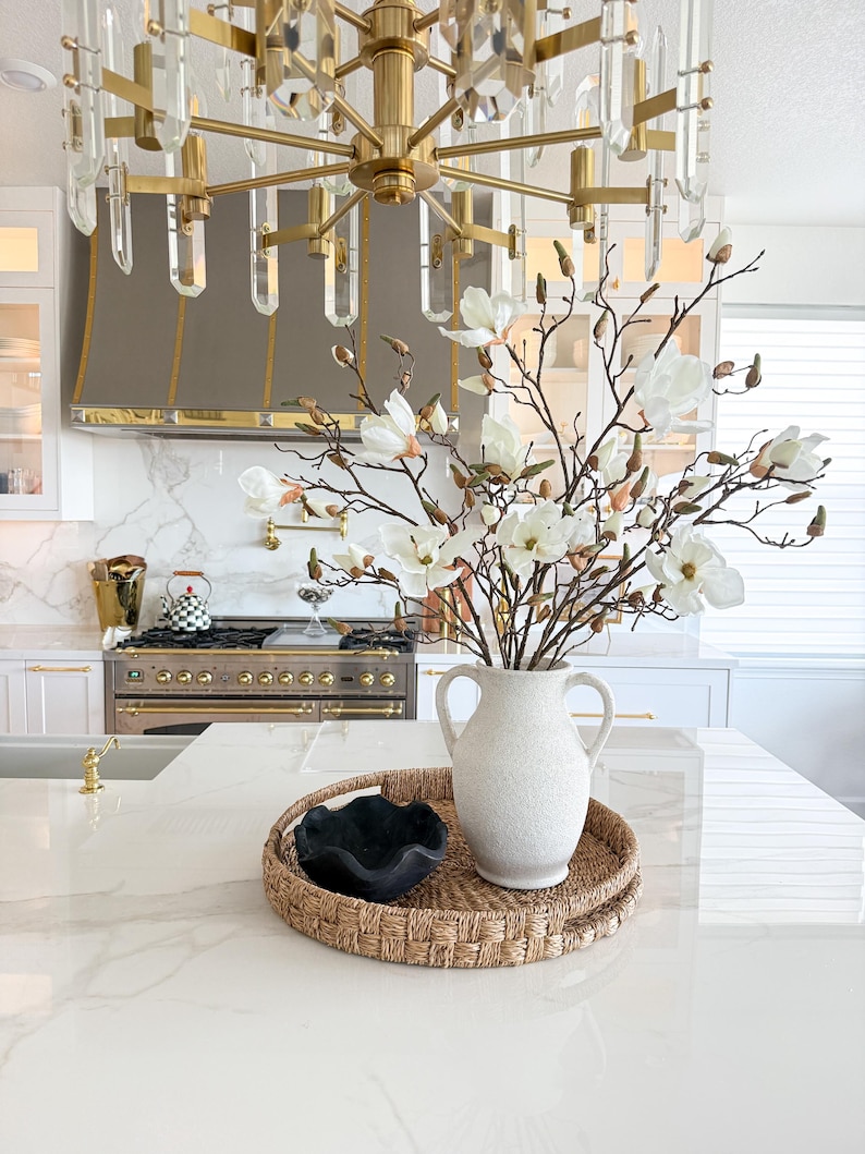 Puede incluir: Una isla de cocina presenta una bandeja tejida con un jarr&oacute;n blanco texturizado lleno de ramas de magnolia blancas. Un cuenco decorativo negro est&aacute; sobre la bandeja. Una l&aacute;mpara de ara&ntilde;a dorada cuelga sobre la isla.