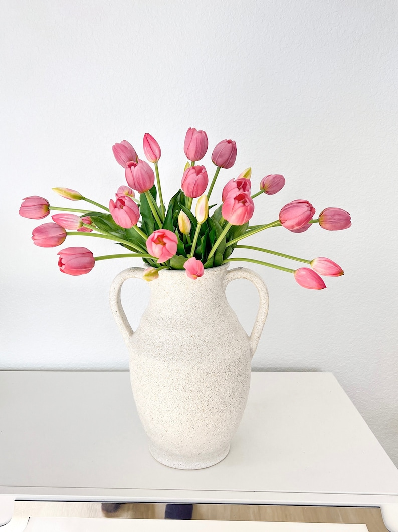 May include: A bouquet of pink tulips in a white ceramic vase with a textured surface. The vase has two handles.