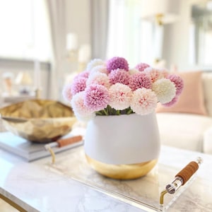 May include: A white vase with a gold rim, filled with a bouquet of pink and white pom pom flowers. The vase is sitting on a clear acrylic tray with a brown leather handle.