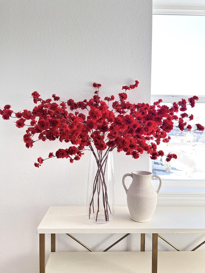 May include: A tall clear glass vase filled with numerous vibrant red flowering branches, positioned next to a smaller, textured off-white ceramic pitcher with two side handles. The arrangement features dense clusters of small, deep red blossoms.