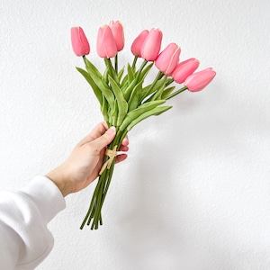 Puede incluir: Un ramo de tulipanes artificiales de color rosa con tallos y hojas verdes, atados con una fina cinta de rafia de color beige. Las cabezas de las flores est&aacute;n cerradas y redondeadas, con una textura suave y un color rosa uniforme.