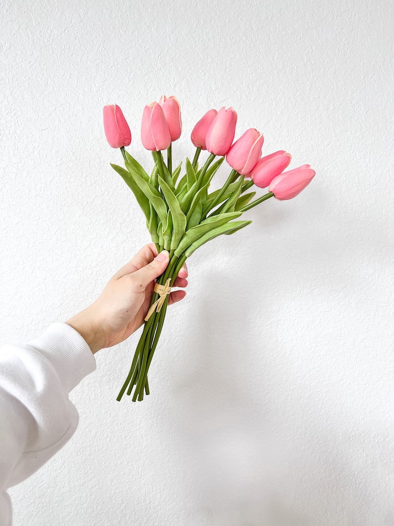 Puede incluir: Un ramo de tulipanes artificiales de color rosa con tallos y hojas verdes, atados con una fina cinta de rafia de color beige. Las cabezas de las flores est&aacute;n cerradas y redondeadas, con una textura suave y un color rosa uniforme.