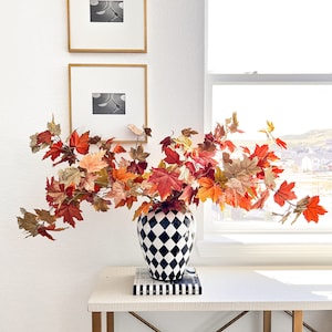 May include: A black and white checkered vase with a large arrangement of fall foliage in shades of red, orange, and gold. The vase is sitting on a white table with gold legs. Two framed black and white photos are on the wall behind the vase.
