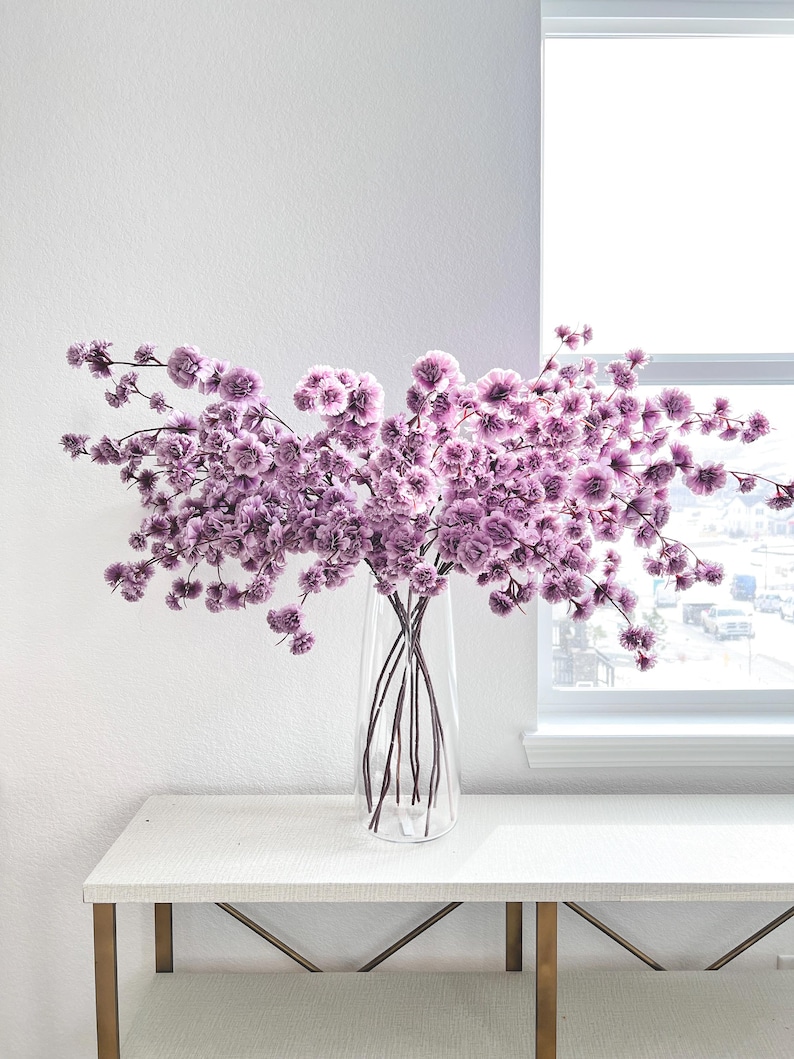 May include: A clear glass vase containing a large, sprawling arrangement of delicate, light purple flowers with numerous small, ruffled blossoms on thin, dark stems, creating a wide, airy display of soft lavender-toned floral textures.