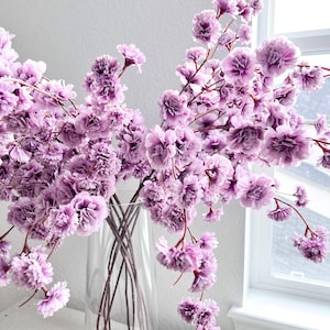 May include: A clear glass vase containing multiple stems of artificial flowers with ruffled, light purple petals. The stems are thin and dark, branching out to support clusters of small, layered blossoms that create a full, airy arrangement.