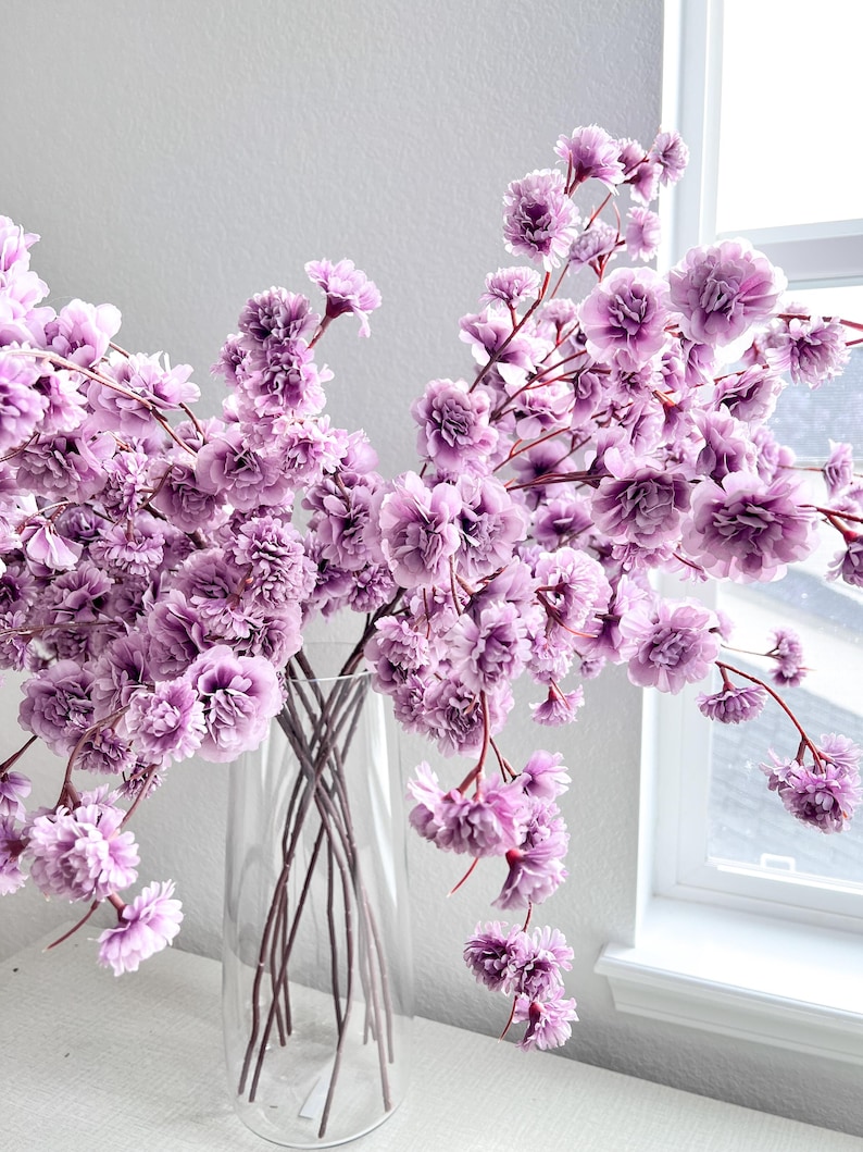 May include: A clear glass vase containing multiple stems of artificial flowers with ruffled, light purple petals. The stems are thin and dark, branching out to support clusters of small, layered blossoms that create a full, airy arrangement.