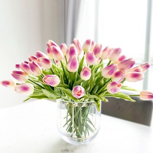 Puede incluir: Un gran ramo de tulipanes artificiales con p&eacute;talos blancos y rosa viol&aacute;ceo y tallos verdes, dispuestos en un florero de vidrio transparente con una estructura de alambre dorado alrededor del exterior del recipiente cil&iacute;ndrico.