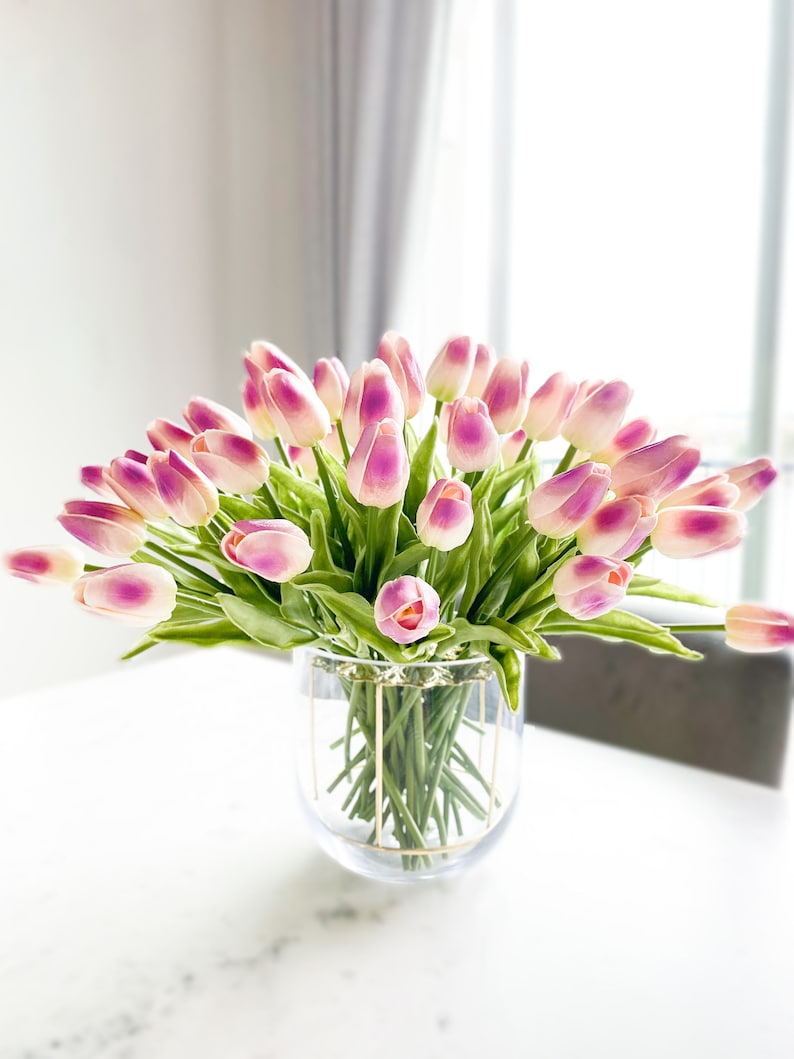 Puede incluir: Un gran ramo de tulipanes artificiales con p&eacute;talos blancos y rosa viol&aacute;ceo y tallos verdes, dispuestos en un florero de vidrio transparente con una estructura de alambre dorado alrededor del exterior del recipiente cil&iacute;ndrico.