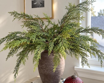 Op de afbeelding: Een donkerbruine, getextureerde, ronde keramische vaas gevuld met weelderige, groene ceder-takken. Naast de vaas staat een ronde, dieprode glazen ornament met een ruitpatroon, rustend naast een zwart-wit gestreept boek.