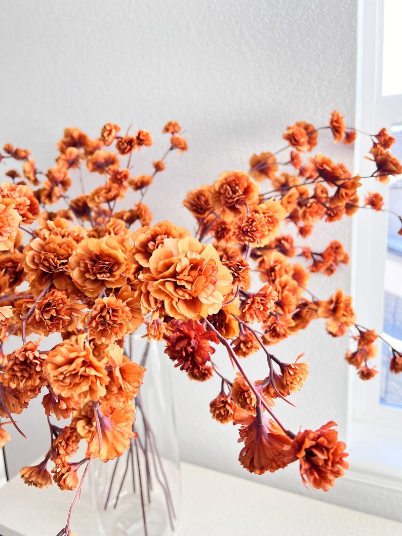 May include: A bouquet of dried orange flowers in a clear glass vase. The flowers are arranged in a loose, cascading style, with the stems extending out from the vase.