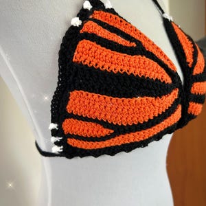Pode incluir: Top de croch&ecirc; feito &agrave; m&atilde;o com um design de asa de borboleta. O top apresenta listras laranja e pretas alternadas, uma borda preta e detalhes brancos. As al&ccedil;as s&atilde;o pretas.