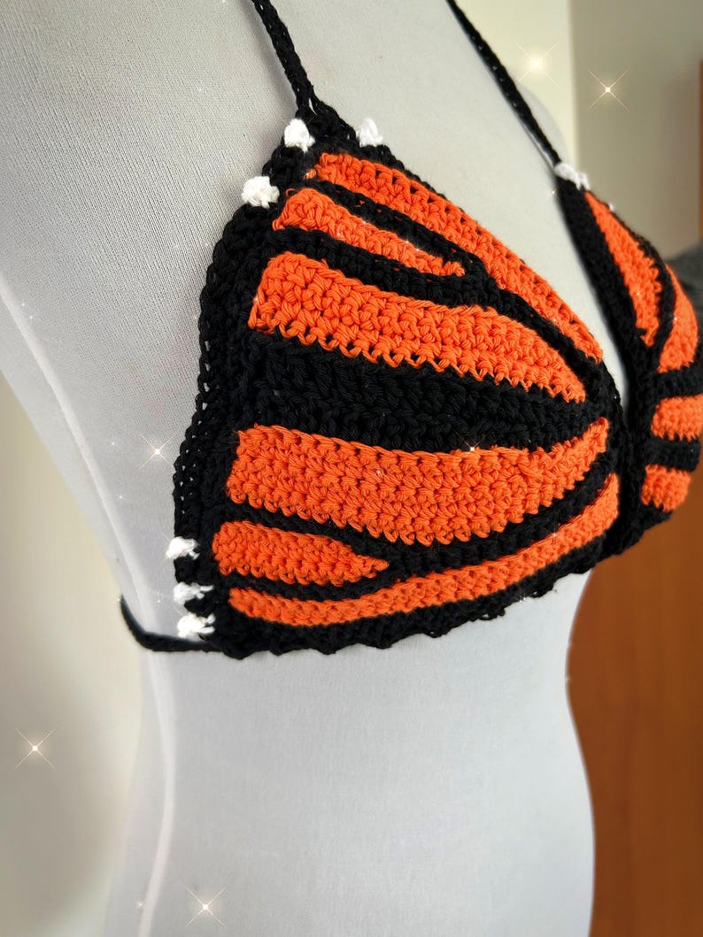 Pode incluir: Top de croch&ecirc; feito &agrave; m&atilde;o com um design de asa de borboleta. O top apresenta listras laranja e pretas alternadas, uma borda preta e detalhes brancos. As al&ccedil;as s&atilde;o pretas.