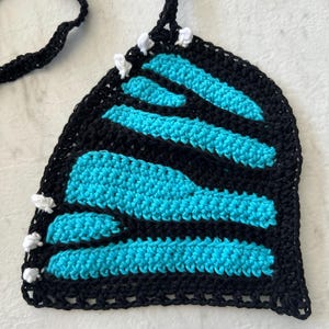 Pode incluir: Top de croch&ecirc; feito &agrave; m&atilde;o com um design de asa de borboleta. O top apresenta uma borda e al&ccedil;as pretas, com listras azul turquesa. Acentos brancos s&atilde;o colocados ao longo das bordas. O top &eacute; feito de fio macio.