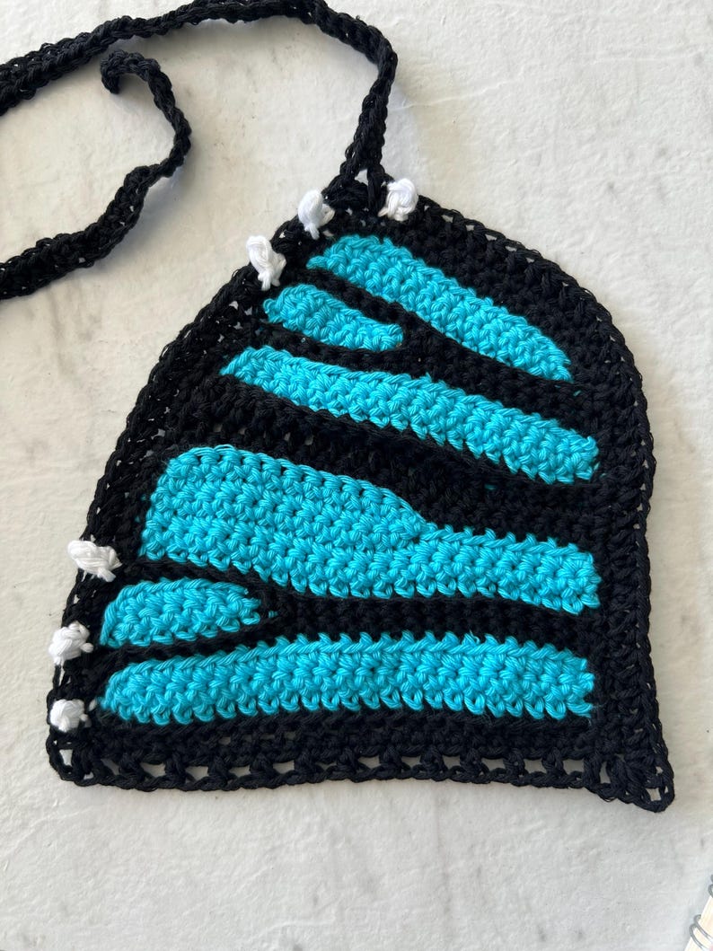 Pode incluir: Top de croch&ecirc; feito &agrave; m&atilde;o com um design de asa de borboleta. O top apresenta uma borda e al&ccedil;as pretas, com listras azul turquesa. Acentos brancos s&atilde;o colocados ao longo das bordas. O top &eacute; feito de fio macio.
