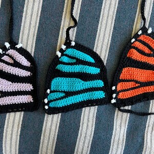 Pode incluir: Tr&ecirc;s tops de borboleta de croch&ecirc; em preto, com asas em lavanda, turquesa e laranja. Cada top tem uma al&ccedil;a preta e detalhes brancos nas bordas, que se assemelham a antenas. Os tops s&atilde;o exibidos em um tecido listrado.