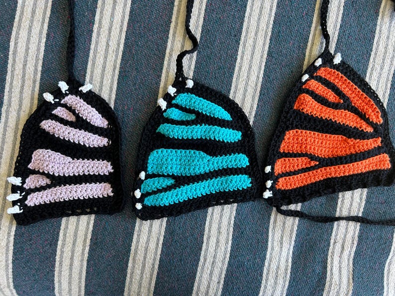 Pode incluir: Tr&ecirc;s tops de borboleta de croch&ecirc; em preto, com asas em lavanda, turquesa e laranja. Cada top tem uma al&ccedil;a preta e detalhes brancos nas bordas, que se assemelham a antenas. Os tops s&atilde;o exibidos em um tecido listrado.